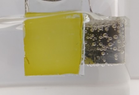 A oxidação da glicerina acontece no fotoanodo de vanadato de bismuto (amarelo) enquanto o hidrogênio (bolhas) é gerado no outro eletrodo (imagem: divulgação/CINE)